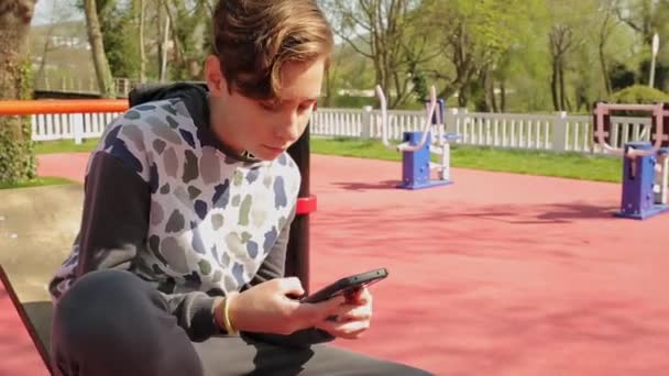 悲しい十代の男の子だけで遊び場に座って スマートフォンを使用しています 屋外だ 現代技術と青少年問題の概念 — ストック動画