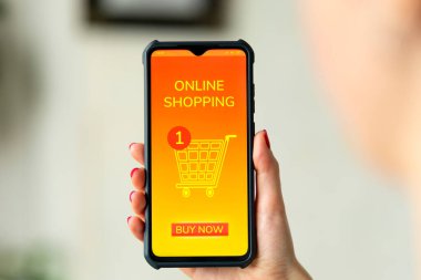 Cihaz ekranında alışveriş arabası olan bir cep telefonunu tutan bir kadının fotoğrafını çek. Arka plan bulanık. Çevrimiçi alışveriş ve tüketim kavramı.