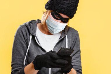 Yeni normal konsept. Siyah şapkalı, eldivenli ve koruyucu maskeli bir kadın hacker elinde bir akıllı telefon tutuyor ve hacklemeye çalışıyor. Sarı arka plan. Siber suç kavramı.
