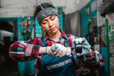 Üniformalı genç bir kadın tamircinin portresi, kafasına eldiven ve güvenlik gözlüğü takıyor, araba parçalarını onarıyor. Kapalı.