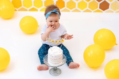 Bir bebek pastanın yanına oturur ve elleriyle bir parça yer. Arka planda sarı bal peteği ve balon tasarımı var. Üst Manzara. İlk doğum günü konsepti.