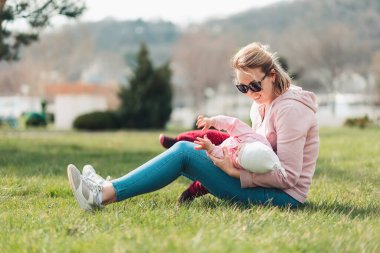 Genç beyaz bir anne çimenlerde oturur ve bebeğine sarılır. Parkta çocuklu bir eğlence. Anneler Günü..