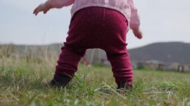 Bebek otların üzerinde çömelip yürümeyi öğreniyor, elleriyle zemini tutuyor. Anne çocuğu alır ve ayaklarının üzerine koyar. Yakın plan, arka plan
