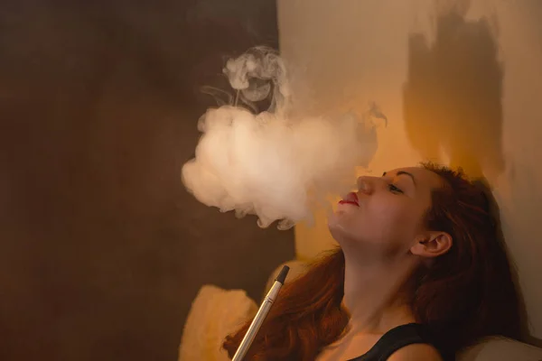 Relaxed on the couch, the woman smokes a hookah and lets out a cloud of steam. Orange color.