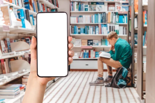 A person holding a mobile phone in hand. In the background boy sitting and reading books in a store, blurred. The concept of online shopping and education