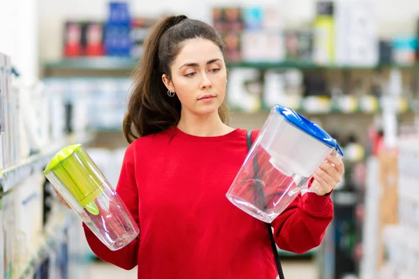 年轻女性的肖像在两家家用电器商店里装有滤水器的卡拉菲之间做出选择 消费主义的概念 — 图库照片