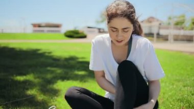 A young Caucasian woman in sports clothes, puts on sneakers and ties her shoelaces. Park or stadium in the background. Concept of sports and healthy lifestyle.