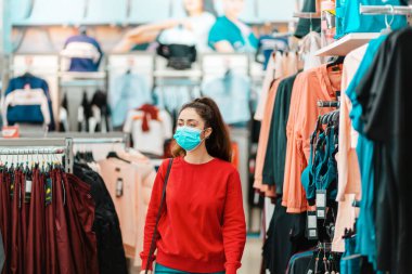 Halka açık yerlerde antiviral koruma. Tıp maskeli genç bir kadının portresi mağazadan kıyafet seçiyor. Virüs salgını sırasında alışveriş kavramı.