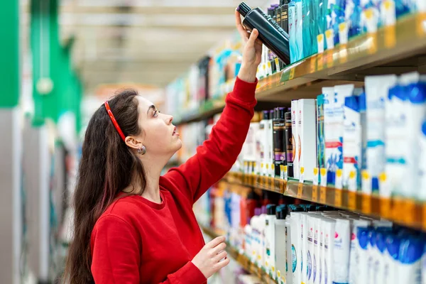 Portrait of a young Caucasian woman in a red sweater, who takes shampoo from the top shelf. Cosmetic shelves are blurred in the background. The concept of buying cosmetics.