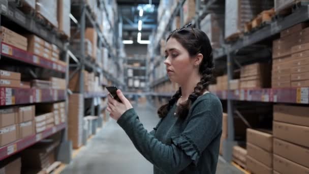 一位年轻妇女用电话检查她的定单 并在仓库的货架上搜寻产品 她抬起头来 对货架的高度感到惊讶 — 图库视频影像