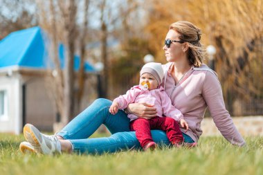 Mutlu bir anne çimenlerde oturur, bebeğine sarılır, bahar güneşinin tadını çıkarır. Çocuklarla açık havada aile eğlencesi.