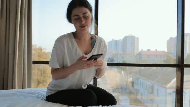 家庭娱乐活动 年轻美丽的女人坐在床上 听音乐 背景中的窗口 — 图库视频影像