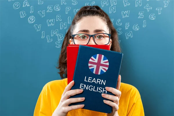 English language day. Portrait of a young woman holding english dictionary, covering half of her face. Blue background with letters. The concept of learning foreign languages.