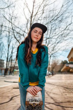 Genç, beyaz bir kadının portresi. Ellerini kaykaya yaslıyor. Arka planda, mavi gökyüzü ve ağaç dalları. Vertikal. Spor yaşam tarzı ve sokak kültürü kavramı..