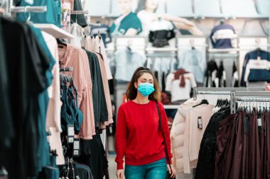 Tüketim. Tıbbi maskeli genç ve güzel bir kadın mağazadan kıyafet seçiyor. Virüs salgını sırasında alışveriş kavramı.