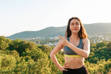 Genç bir atletik kadın eliyle bir şeyi işaret ediyor ve kameraya bakıyor. Yoga, spor ve meditasyon konsepti. Arka planda köyün ve dağların manzarası. Boşluğu kopyala.