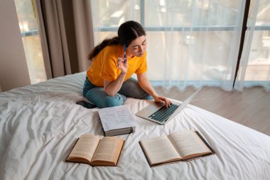 Online eğitim. Gündelik giysiler içinde bir kadın bir yatakta oturuyor ve bir dizüstü bilgisayar üzerinde çalışıyor. Kitaplar yan yana yatıyor. Çevrimiçi kurslar kavramı.