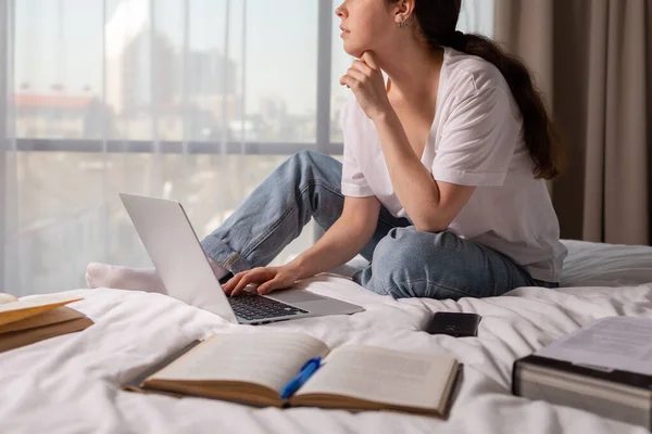 A woman sits in bed and works on a laptop. Books and a smartphone are scattered on the bed. Close up. The concept of online courses and online education.