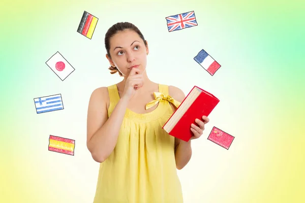 English language Day. A young thoughtful woman in a yellow dress holds a book in her hands, choosing a language to study. Yellow-green background with flags of different countries. Concept of learning foreign languages