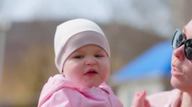 Sevimli bir bebek gülümser ve annesinin etrafında istikrarsız bir şekilde yürür. Bir anne çocuğunu öper ve ona sarılır. Yakın plan portre. Alt köşeden bak. Parkta eğlence..