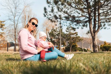 Gülümseyen bir anne çimenlerde oturup bebeğine sarılır. Çocuklarla açık hava eğlencesi.