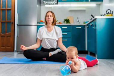 Genç beyaz bir anne, bebeği yakınlarda yatıp oyuncaklarla oynarken mutfakta meditasyon yapıyor. Çocuklarla yoga kavramı.