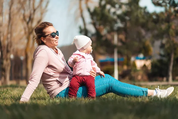 一位笑着的母亲坐在草地上 抱着她的孩子 享受着春天的阳光 有子女的家庭户外运动 — 图库照片