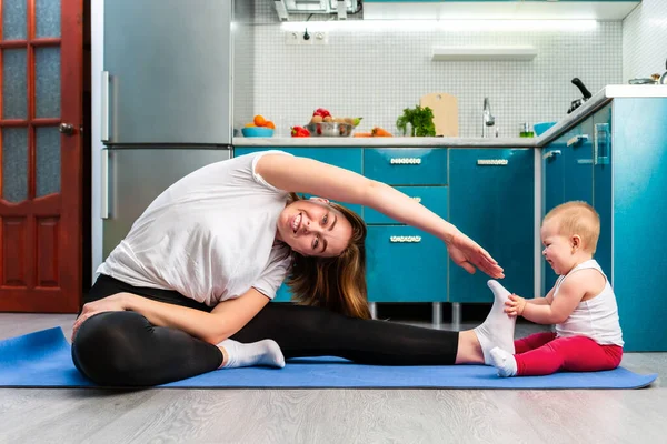 Bir anne ve bebeği mutfakta bir paspasın üzerinde geriniyorlar. Çocuk annenin bacağının yanına oturur ve elleriyle onu tutar. Çocuklarla evde spor eğitimi kavramı.