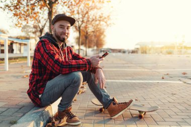 Kaykay yapmak. Sakallı, şapkalı, şık elbiseli, kaldırımda elinde akıllı bir telefonla oturan yakışıklı bir adam. Ayaklarının altında bir kaykay yatıyor. Arka planda, şehrin boş bir caddesi. Boşluğu kopyala.