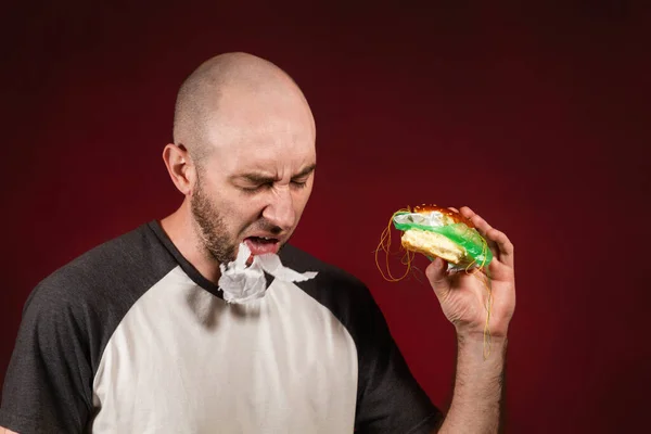 The concept of fast food and ecology. A bald man with a beard, holding a hamburger and spitting plastic filling. Burgundy background.