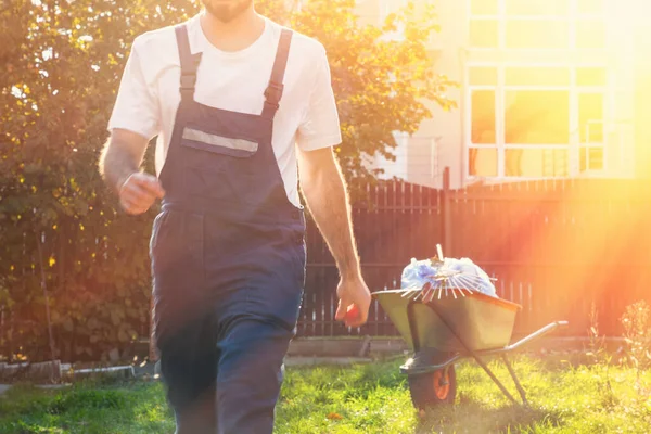 Gün batımında mavi üniformalı bir adamın yakın çekimi, hareket halinde, bahçıvan işi bitirdi. El arabası çantalar ve arka planda tırmıkla gezdi..