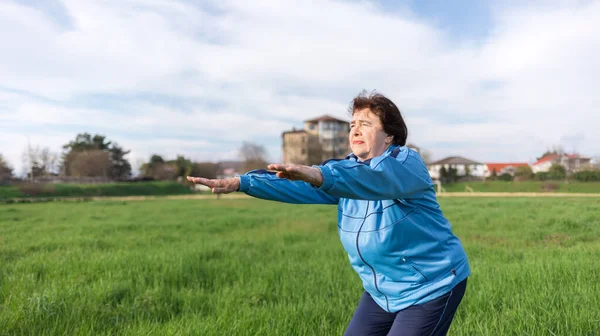 Concept of a healthy lifestyle and sports. Smiling elderly woman in sportswear performs a squat exercise in the park. Grass in the background. International Day of Older Persons.