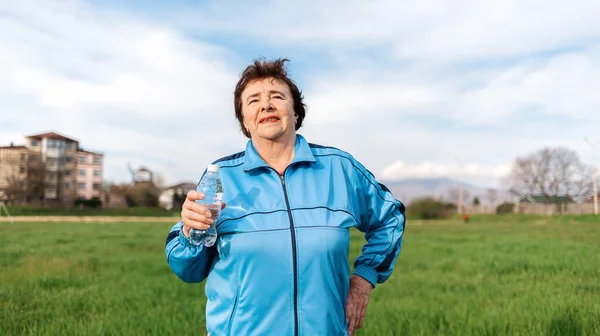 The concept of the International Day of Older Persons. Portrait of an elderly smiling woman in sports clothes, holding a bottle of water. Outdoor activities.