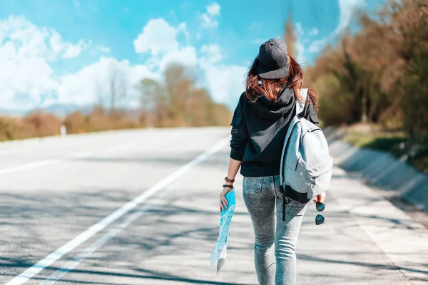The concept of hitchhiking trips and local travel. Hipster woman in a cap, with backpack walking down the road holding a paper map in her hand. Blurred road on the background. Rear view.