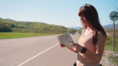 Yolun kenarında duran ve kağıt bir haritaya bakan genç bir kadın gezginin portresi. Yerel seyahat ve otostop konsepti