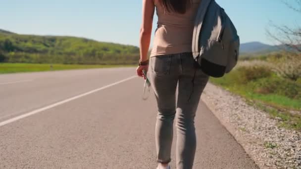 En kvinna med ryggsäck går längs vägen. Bakifrån. Flyttar ner kameran. Begreppet liftresor — Stockvideo
