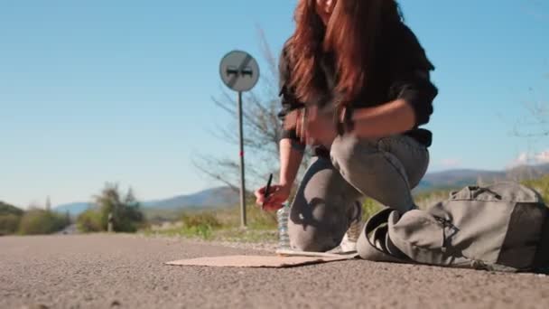 Sommarsemester. Kaukasiska unga kvinna skriva på en kartong skylt, sitter på landsbygden väg väntar på en bil. Begreppet liftresor och lokala resor — Stockvideo