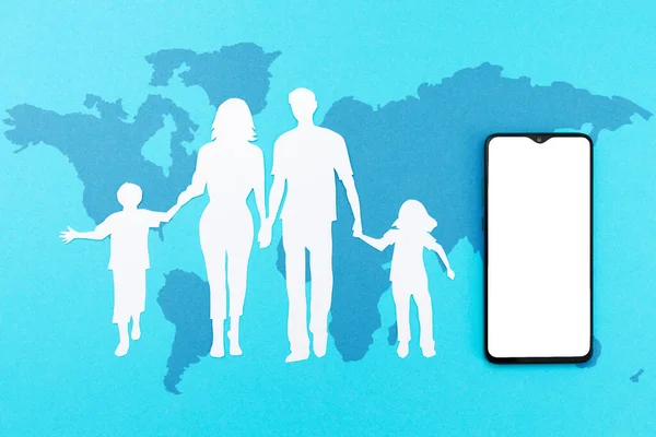 空のスクリーンと世界地図で青の背景に紙から切り取られた家族のシルエットを持つ携帯電話 平置きだ モックアップ 世界的な予防接種の概念 — ストック写真