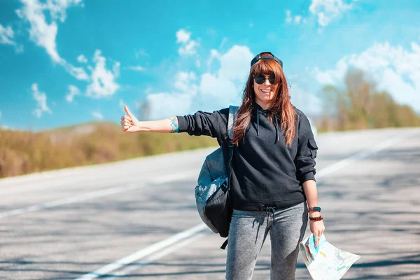 Concept of hitchhiking and local travel. A hipster happy girl in cap and sunglasses with tattoed hand stand along the road with a paper map and hitchhiking with thumbs up. Copy space.