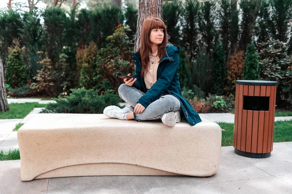若い美しい女性が公園のベンチに座って スマートフォンを使用しています — ストック写真
