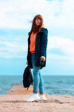 Elinde sırt çantası olan ceketli genç bir kadın. Arka planda, mavi deniz ve bulutlu gökyüzü. Dikey. Uzayı kopyala Özgürlük ve seyahat kavramı.