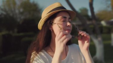 Yaz zamanı. Hasır şapkalı, güneş gözlüğünü çıkarıp kameraya bakan genç bir kadının portresi. Arka planda gün batımı. Çekim.