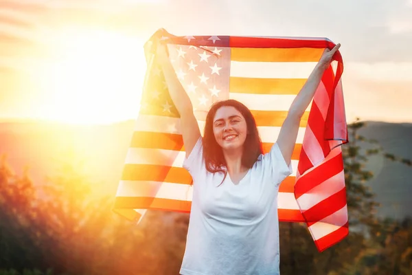 独立日 一个快乐的高加索年轻女人手里拿着一面美国国旗 举过头顶 夕阳西下美国国定假日的概念 — 图库照片