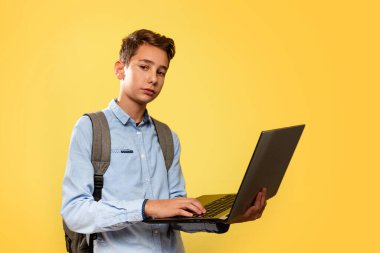 Modern teknoloji ve eğitim kavramı. Mavi gömlekli, sırt çantalı, elinde dizüstü bilgisayar olan beyaz bir çocuk. Sarı arka plan. Boşluğu kopyala ve kapat.