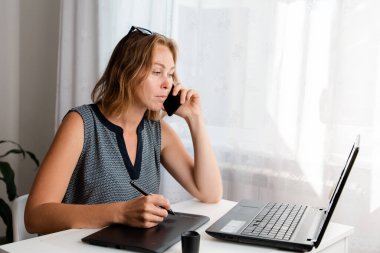 Uzaktan kumanda. Bir kadın evde grafik tabletle çalışıyor ve telefonda konuşuyor. Karantina, serbest çalışma ve kendini izole etme kavramı..
