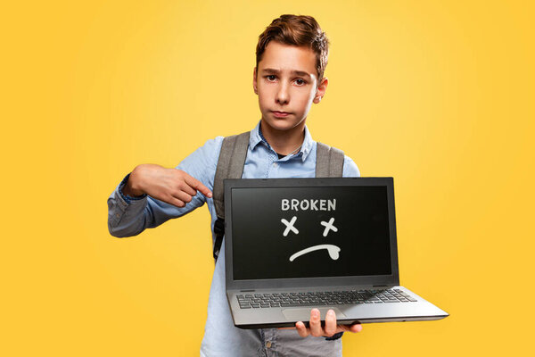 Concept of modern technologies and education. A disgruntled Caucasian teenage boy holds an open laptop and points at it with his finger. Yellow background. Copy space. The inscription Broken.
