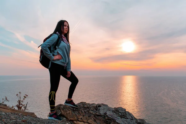 The concept of Hiking and sports recreation. A woman with a backpack on her back poses against the sea and sky. Sunset. Copy space.