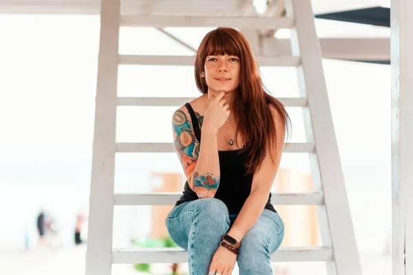 若い笑みを浮かべて美しい女性のタトゥーを彼女の腕の上に ポーズを 白い階段に座っている — ストック写真