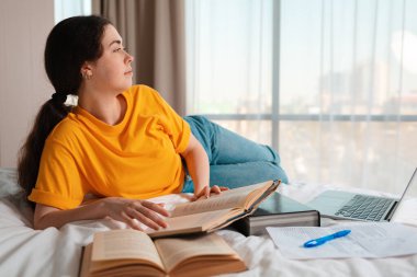 Genç beyaz bir kadının portresi yatağında uzanıyor ve elinde bir kitap tutuyor. Laptop ve belgeler yan yana duruyor. Online kurslar ve çevrimiçi eğitim kavramı.