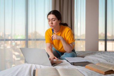 Güzel bir kadının portresi bir yatakta oturup dizüstü bilgisayarla çalışmaktır. Kitaplar yan yana yatıyor. Çevrimiçi kurslar kavramı.